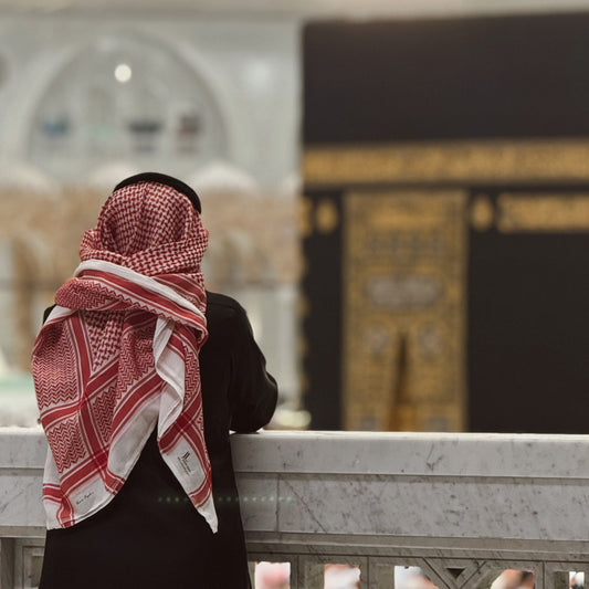 man wearing black saudi collared thobe with red and white  shemagh while looking at the kaaba