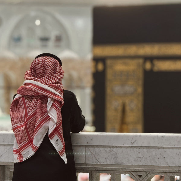 man wearing black saudi collared thobe with red and white  shemagh while looking at the kaaba