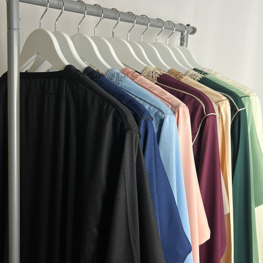 A row of colourful men’s thobes hanging on white wooden hangers, including black, navy, light blue, pink, maroon, cream and green styles, displayed on a clothing rail in soft studio lighting.