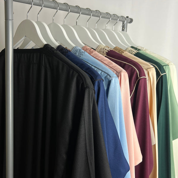 A row of colourful men’s thobes hanging on white wooden hangers, including black, navy, light blue, pink, maroon, cream and green styles, displayed on a clothing rail in soft studio lighting.